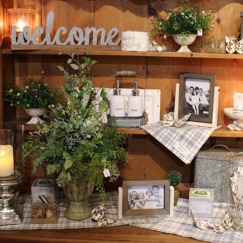 A closeup of a shelf unit with a variety of home decor items including: galvanized welcome sign, floral arrangements, soap and lotion set, picture frames, salt and pepper shakers and more.