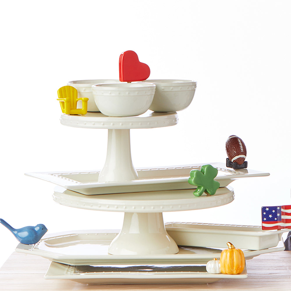 A stack of white ceramic serving dishes and trays, each with a colorful sculptural attachment celebrating a different occasion or season.  Attachments featured include heart, Adirondack chair, football, clover, bird, flag, and pumpkin.