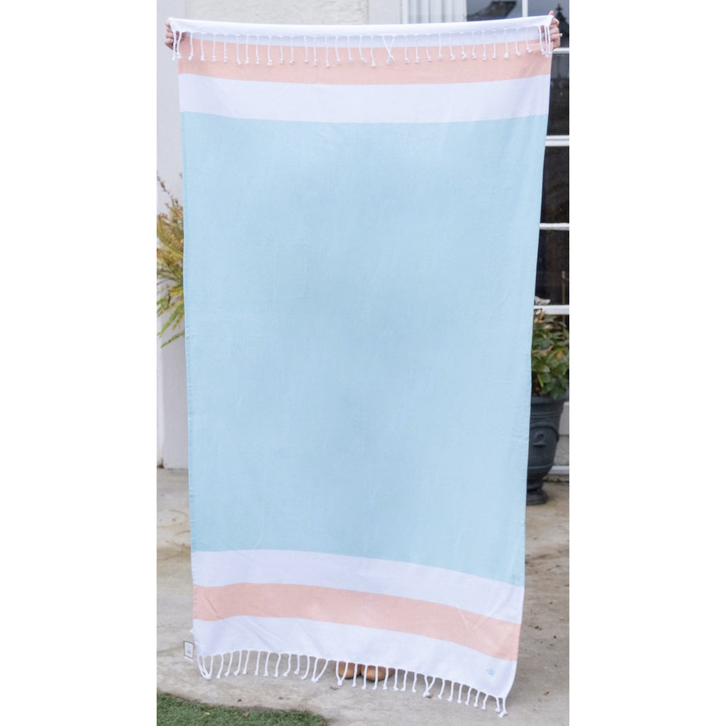 A turquoise blue beach towel with stripes at the top and bottom.  The stripes are wide and horizontal, white, orange white.  There is white fringe along the top and bottom of the towel.
