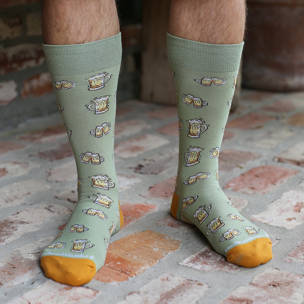 A photo of a man's legs from below the knee, showcasing a pair of sage green dress socks with graphics of beer mugs on them.  The toe and heel is an amber gold.