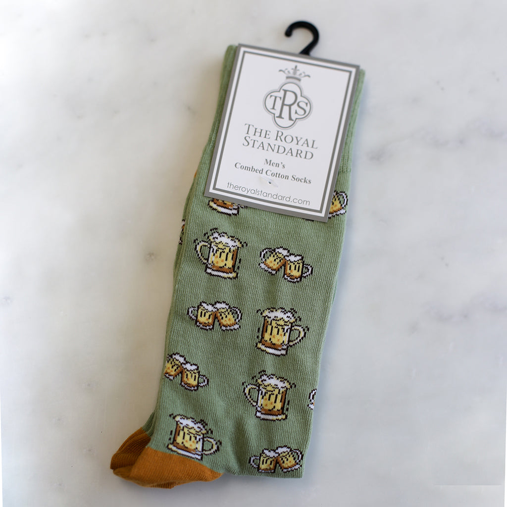 A photo of mens dress socks in their packaging.  The socks are sage green with images of beer mugs.  The background is white marble.  A large tag on the socks reads 'TRS The Royal Standard Men's Combed Cotton Socks - TheRoyalStandard.com'