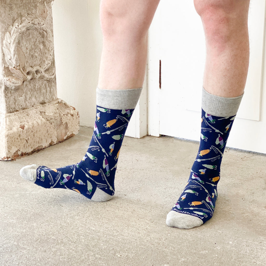 A photo of a man's legs from below the knee, showcasing a pair of navy blue dress socks with graphics of fishing rods and lures on them.  The toe and heel is gray.