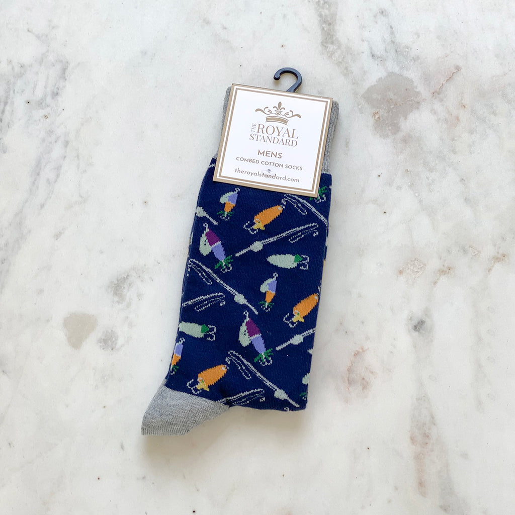 A photo of mens dress socks in their packaging.  The socks are navy blue with images of fishing reels and lures.  The background is white marble.  A large tag on the socks reads 'TRS The Royal Standard Men's Combed Cotton Socks - TheRoyalStandard.com'