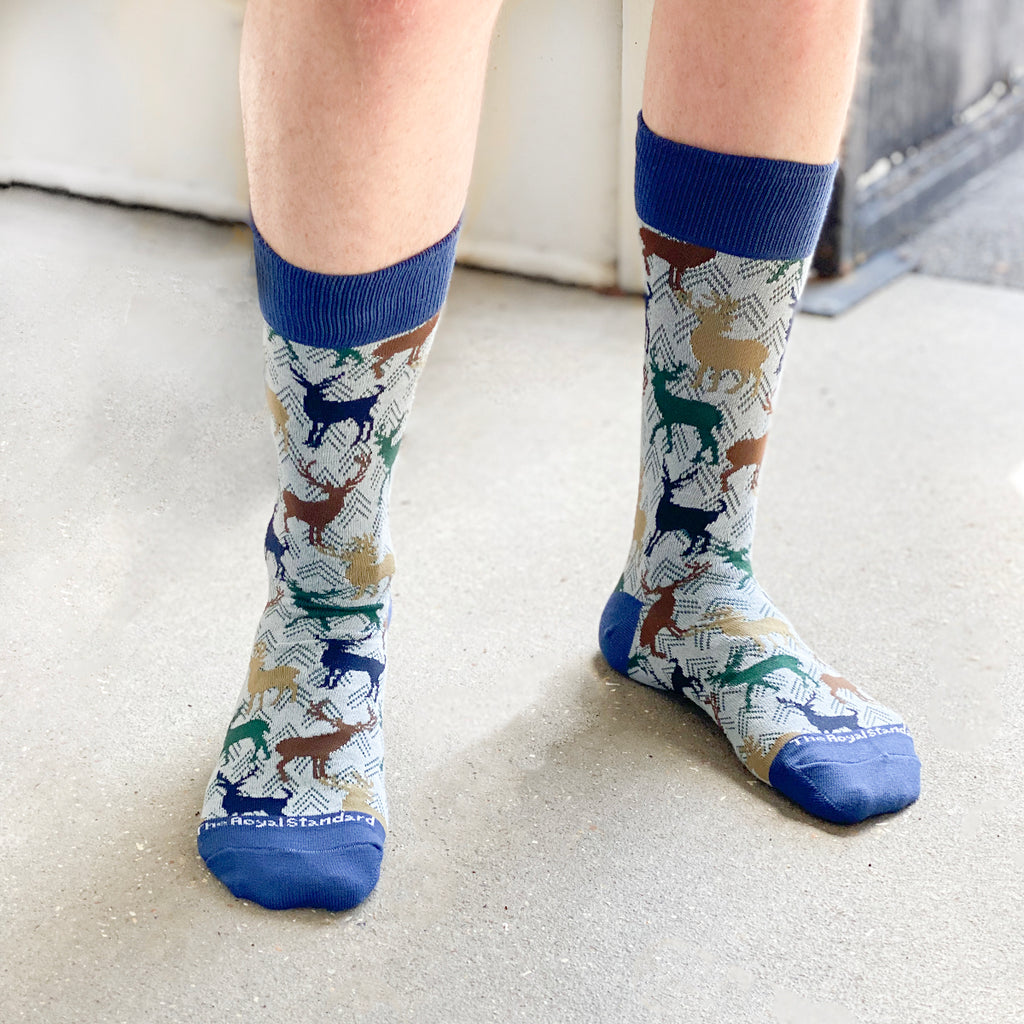 A photo of a man's legs from below the knee, showcasing a pair of light blue dress socks with graphics of silhouettes of stags in tan, blue, and green.  The toe and heel is a cobalt blue.