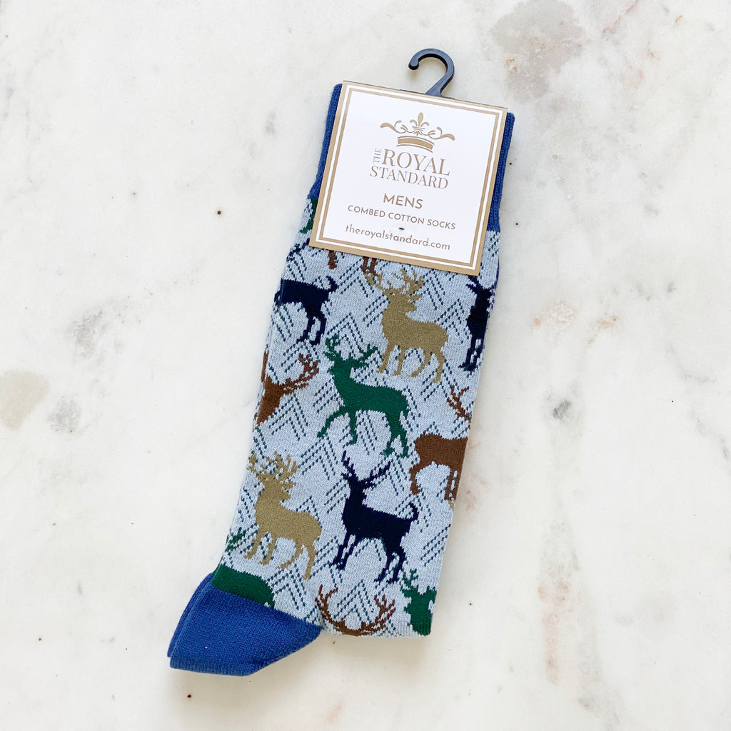 A photo of mens dress socks in their packaging.  The socks are light blue with images of tan, green, and black silhouettes of stags.  The background is white marble.  A large tag on the socks reads 'TRS The Royal Standard Men's Combed Cotton Socks - TheRoyalStandard.com'