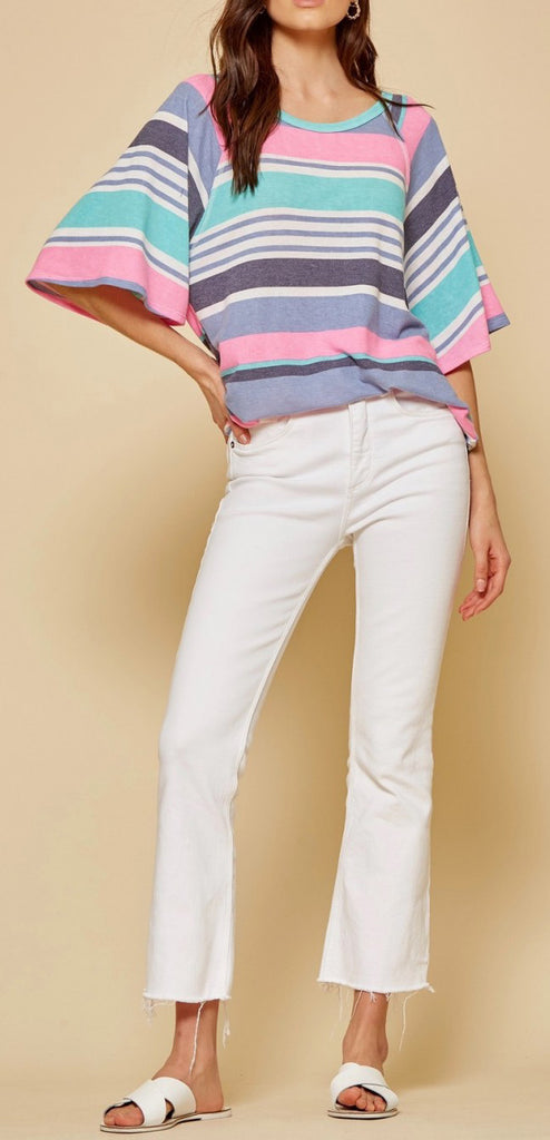 Front view of model wearing a slightly oversized striped top with white jeans and white sandals.  Top has alternating thin and thick stripes in pink, teal, gray, purple and white.