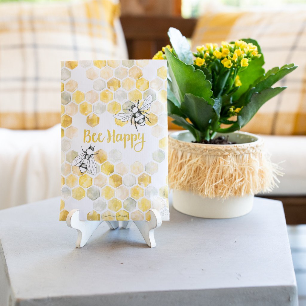 A sachet on an easel on top of a gray table.  Behind the sachet is a flower in a pot with tassel trim.  The sachet has an image of hexagons linked together like a hive with two bees on it, and the text 'Bee Happy'