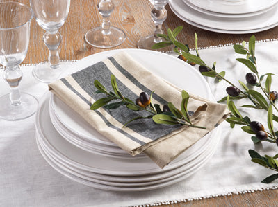 A place setting on a wooden table with a stack of white plates on a white runner.  On top of the plates is a folded tan napkin with a thick gray stripe and a thin gray stripe on either side of it.  On top of the napkin is a decorative olive branch.