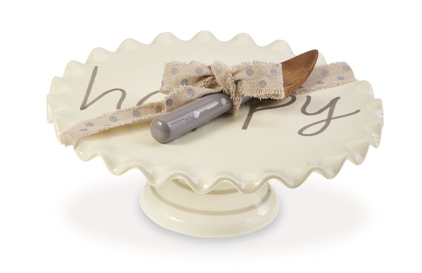 White ceramic cake pedestal that says 'happy' on it in gray text.  A coordinating serving knife with a wooden finish and a gray handle with polka dots is tied to the pedestal with polka dotted ribbon.
