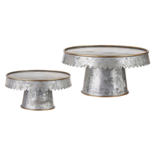 A set of galvanized pedestal stands, one is larger than the other.  Both feature a gold rim around the top of the pedestal, and around the bottom of the base.  The edge of the top of the pedestal curves down and has a scalloped cut detail.