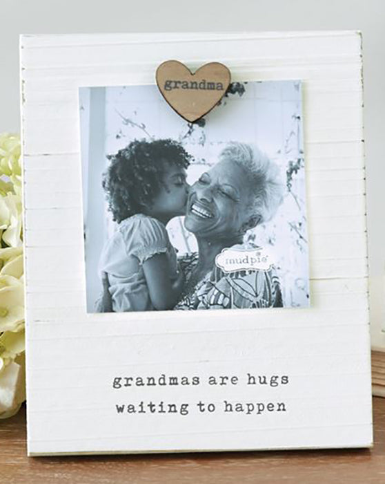 White wooden board with a heart shaped magnet that says 'grandma' on it.  The magnet holds a photo of a granddaughter and her grandmother above a printed saying 'grandmas are hugs waiting to happen'.  Frame is on a wooden table in front of a white wall and a gardenia flower.