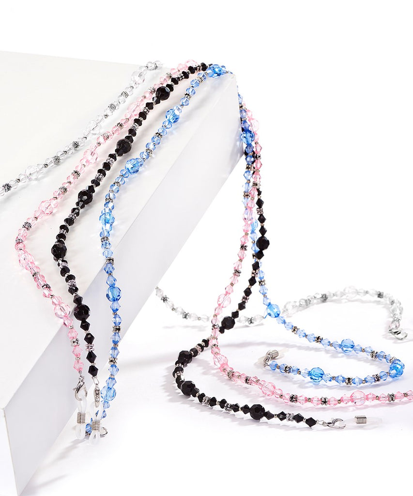 A collection of 4 glass beaded lanyards on a white background draped over a white angled set piece.  From left, there is clear, pink, black, and blue.  Each lanyard cascades off the set piece and collects on a lower level showcasing the lanyards flexibility and facets of the crystals.