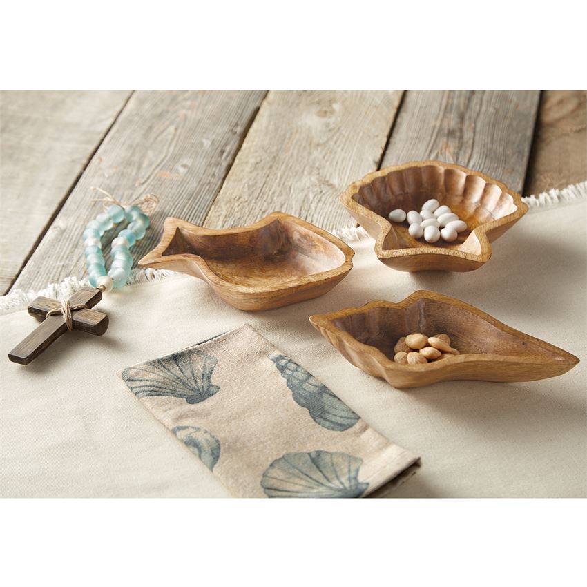 A collection of wooden bowls, one shaped like a fish, a fan shell, and a conch shell, next to a cross with sea glass beads and a napkin with watercolor shells on it