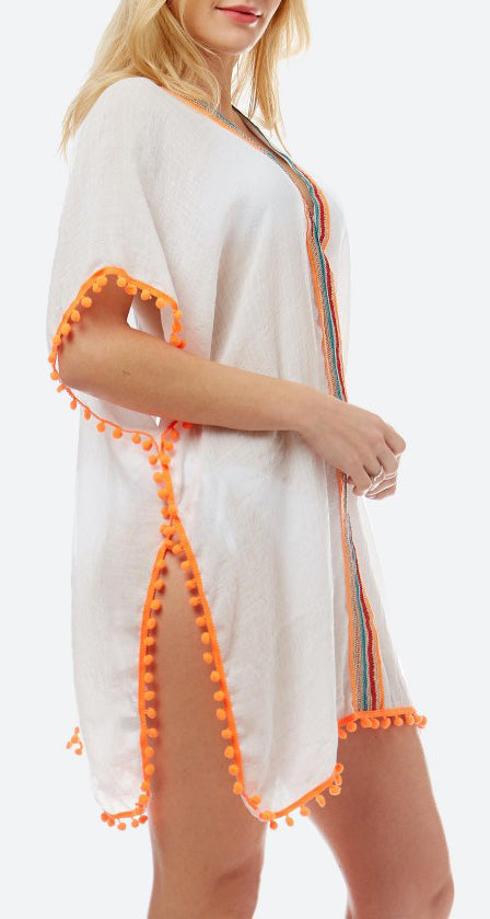 View of the side of a model wearing a loose fitting white top with turquoise, gold, red and orange stripes around the neck and down the middle.  The bottom and arm are accented with orange pom pom trim.