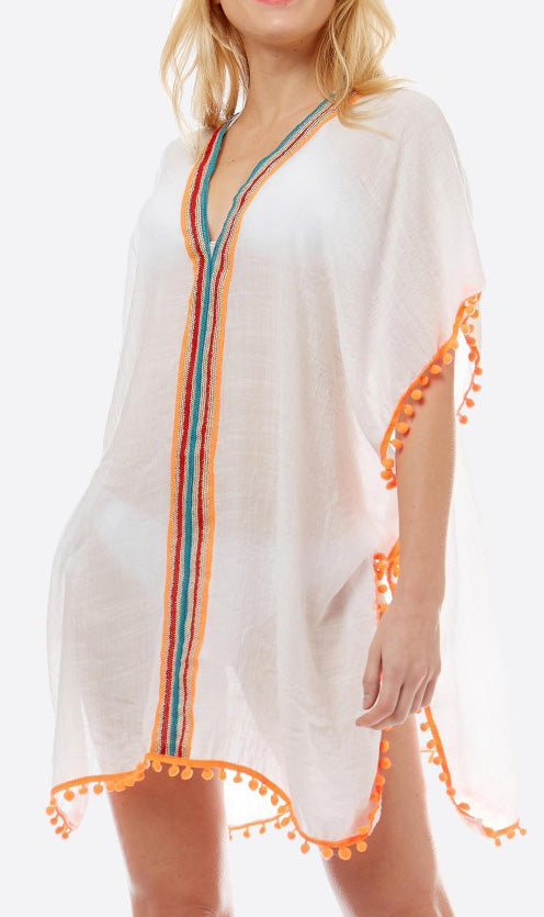 View of the front of a model wearing a loose fitting white top with turquoise, gold, red and orange stripes around the neck and down the middle.  The bottom and arm are accented with orange pom pom trim.