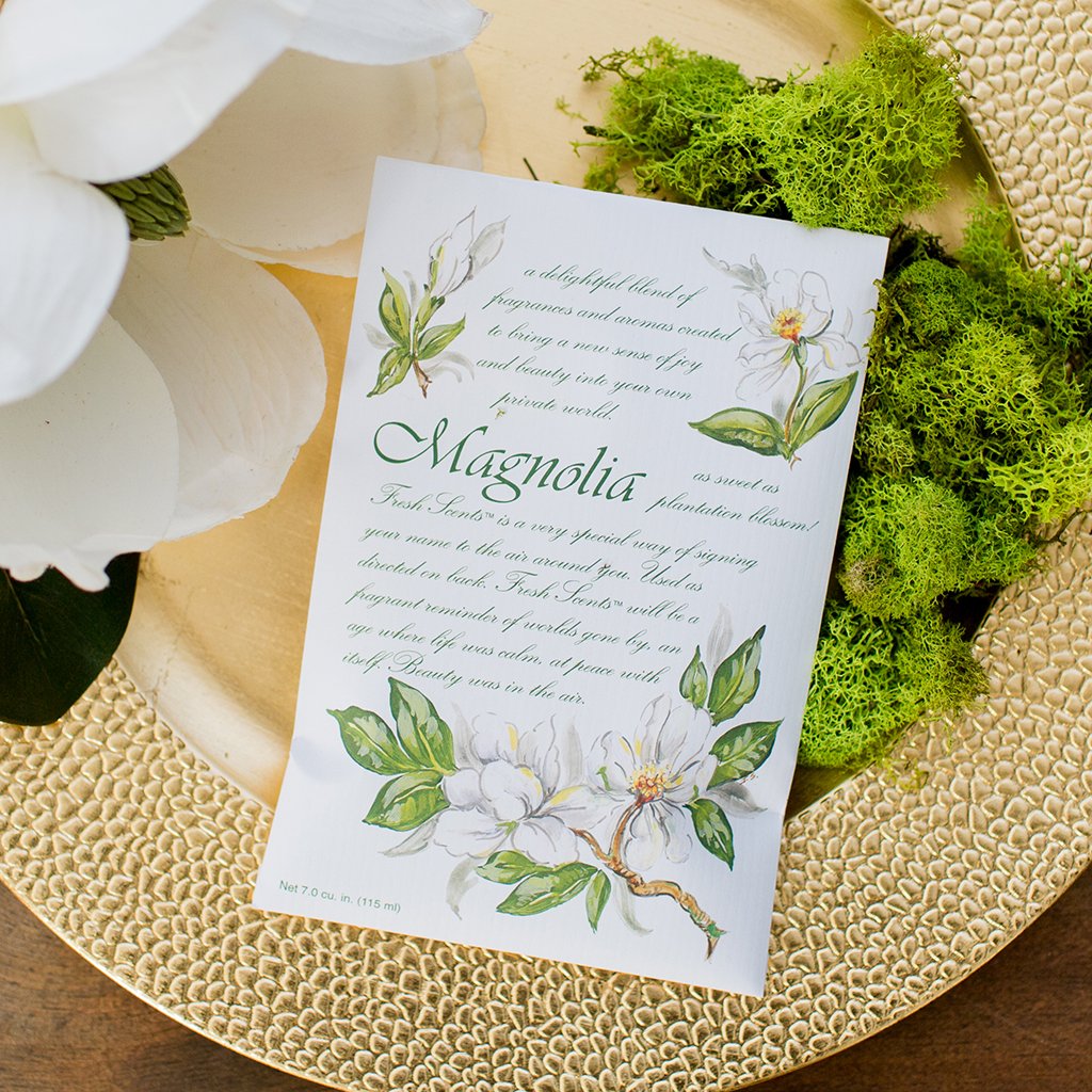 An image of a sachet packet on a gold bowl with moss and a large magnolia flower to the left.  The cover of the packet has text describing the scent in the middle of illustrations of magnolia blossoms.