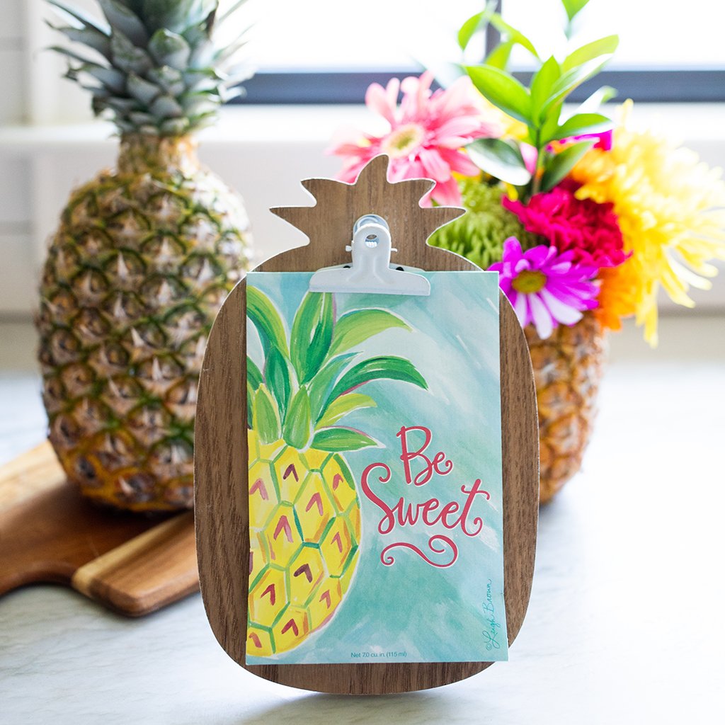 A photo of a sachet packet clipped to a wooden pineapple shaped board.  Behind the board is a pineapple on a cutting board, and a pineapple being used as a vase for a colorful bouquet.  The sachet packet itself showcases a painted pineapple on a blue background and the text 'be sweet'