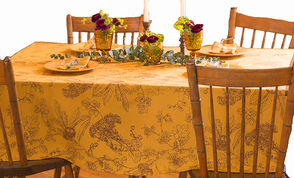 Table set for four with amber glasses with flowers in them, candlesticks and yellow plates.  A sprig of eucalyptus is in the center of the table.  The table cloth has a gold background and burgundy line work of fall leaves and flowers.