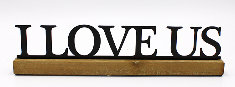 Metal text sign that states 'I Love Us' in black letters with serif.  Letters are attached to a wooden board below.  Sign is in front of a white background.