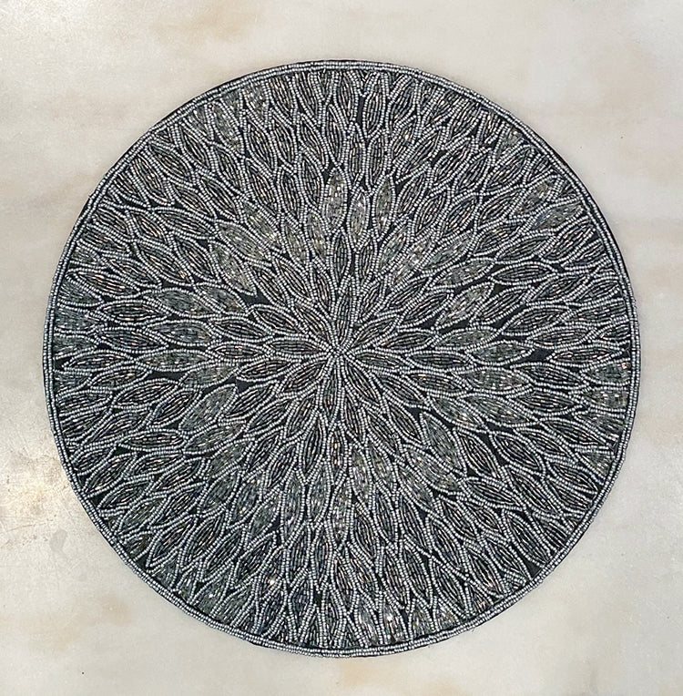 View from above of a glass beaded placemat with a floral pattern.  Beads are in shades of gray and silver.  Round placemat sits on top of a marble surface.