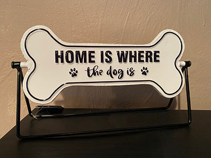 White enamel bone shaped sign on a black metal stand.  Sign can rotate to show a different saying.  Saying on this side is 'Home is where the dog is'