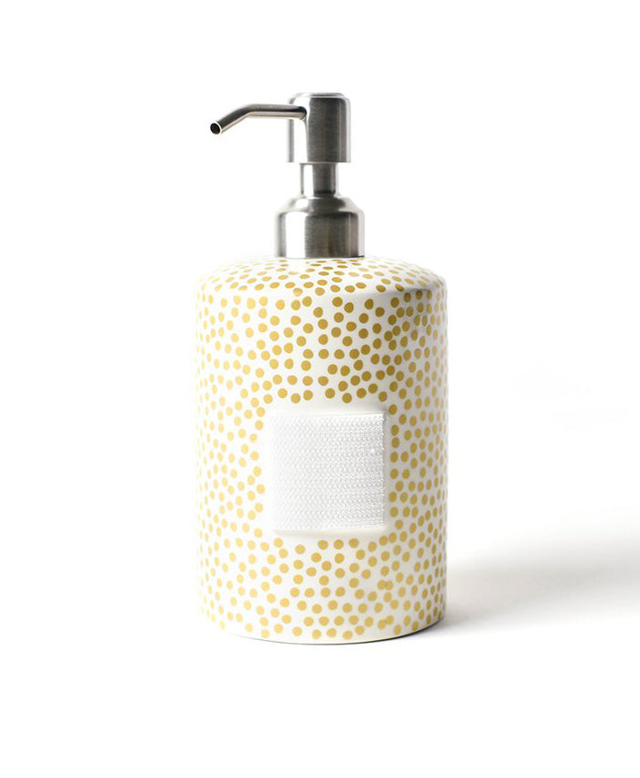 A hand pump soap dispenser on a white background.  The pump itself is polished chrome and the body of the container is a white cylinder with a random gold polka dot pattern.  The front of the cylinder has a rectangular square  piece of velcro for decorative attachments.