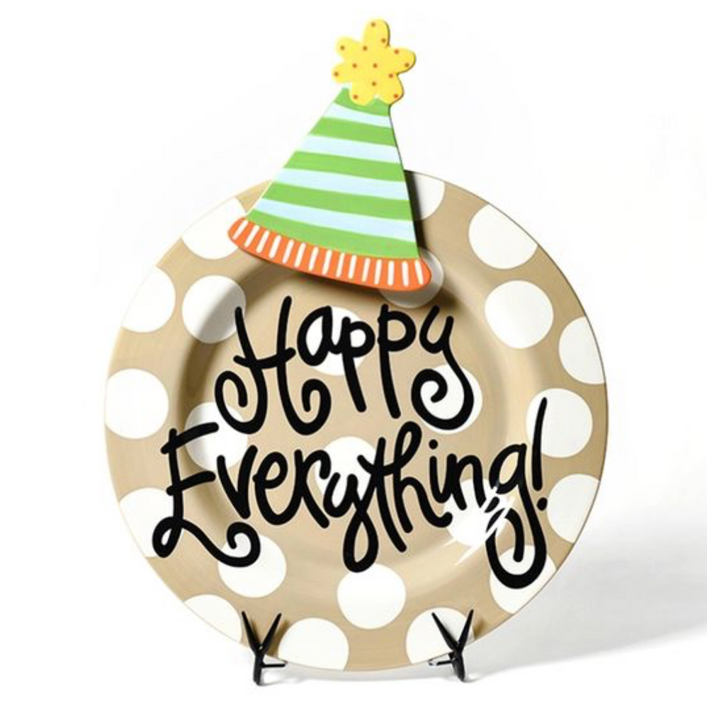 A round platter on a black plate stand in front of a white background.  The plate itself is tan with white polka dots, and has black text in a fun font over the entire platter that says 'Happy Everything!'.  At the top center of the platter is a ceramic attachment in the shape of a conical party hat with a pom on top.  The hat is green and light blue striped with an orange and yellow vertical stripe rim.  The pom is yellow with red polka dots.