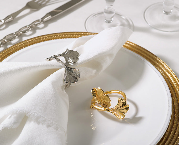 A place setting on a white table cloth.  On top of a gold charger is a white plate.  On the white plate is a napkin with a silver napkin ring with two leaves on it inspired by the ginko leaf.  Next to the napkin is the same napkin ring in gold.