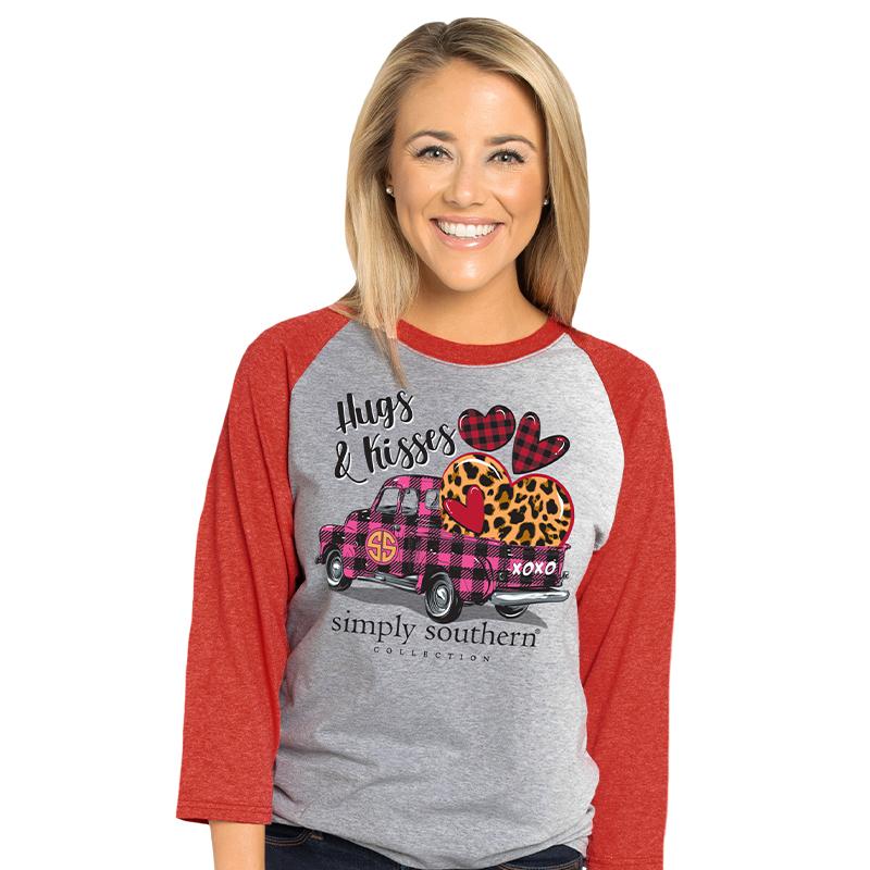 Model wearing gray t-shirt with red long sleeves.  Image printed on shirt is of a pink and black plaid truck with a variety of plaid and leopard hearts in the back.  Text above the truck says 'Hugs and Kisses'.  Text below the truck says 'simply southern collection'