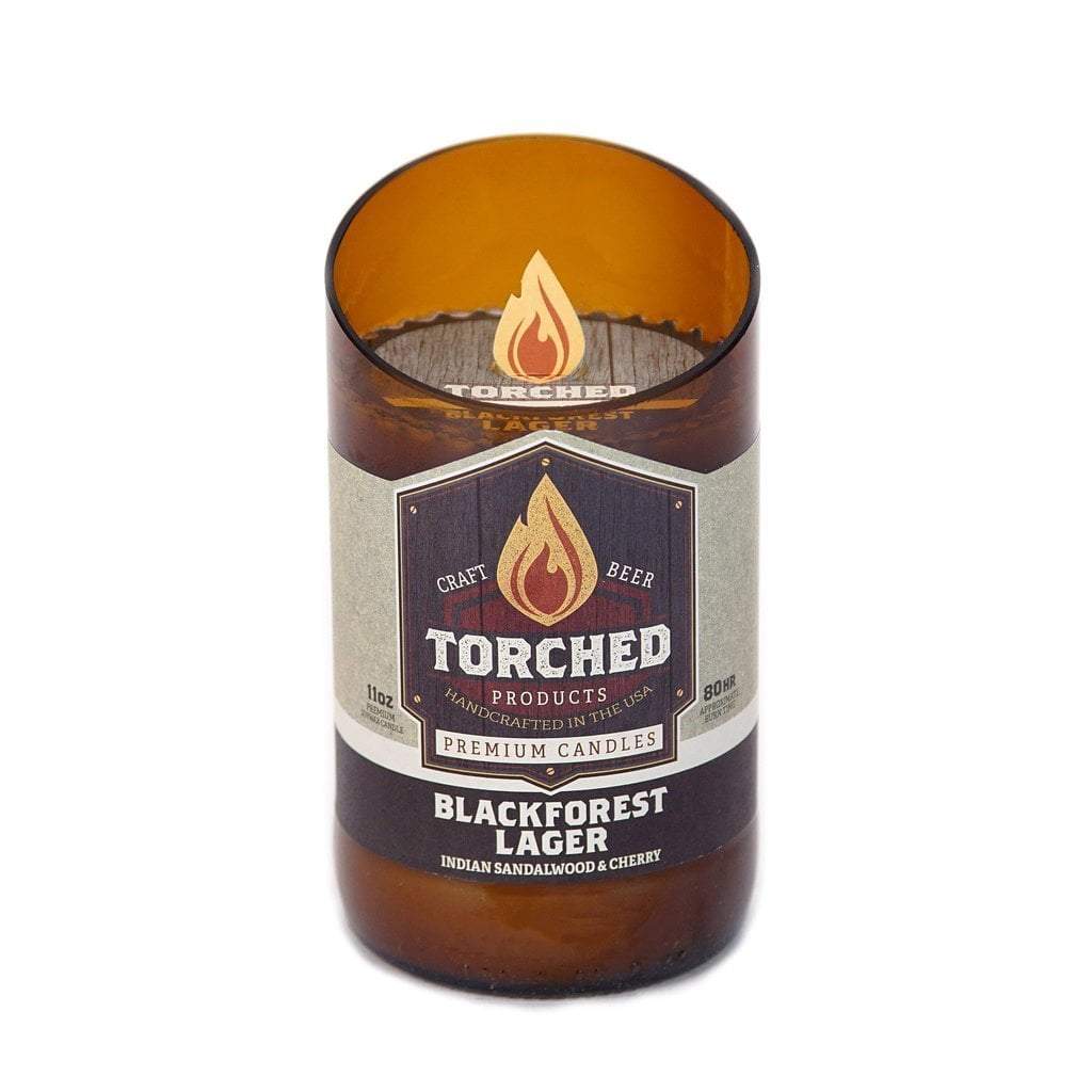 An 11 oz. beer bottle with the top cut off at a 20 degree angle.  The bottle has been filled with wax and is being sold as a candle.  The label shows an illustration of a flame and says 'Torched, craft beer premium candles.  Blackforest Lager, Indian Sandlewood & Cherry'