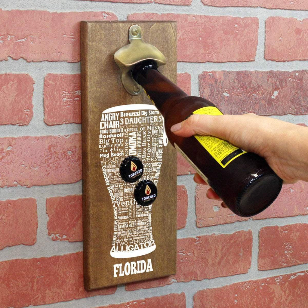 An image of the wall mounted beer bottle opener, with pilsner word art, mounted to a brick wall.  A bottle is being held under the opener, and there are two bottle caps magnetized to the word art.