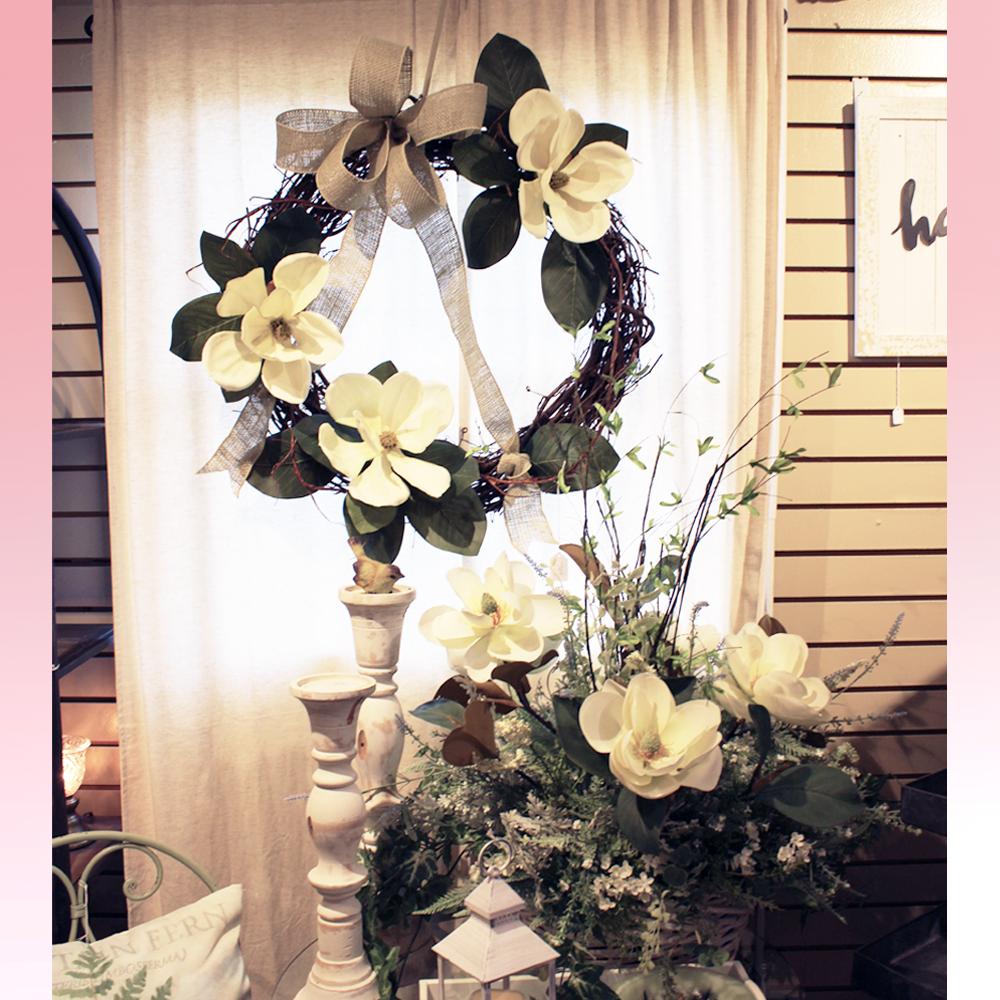 A Magnolia wreath with bow hung behind an accompanying table top arrangement.  Next to the arrangement are two tall wooden candlesticks