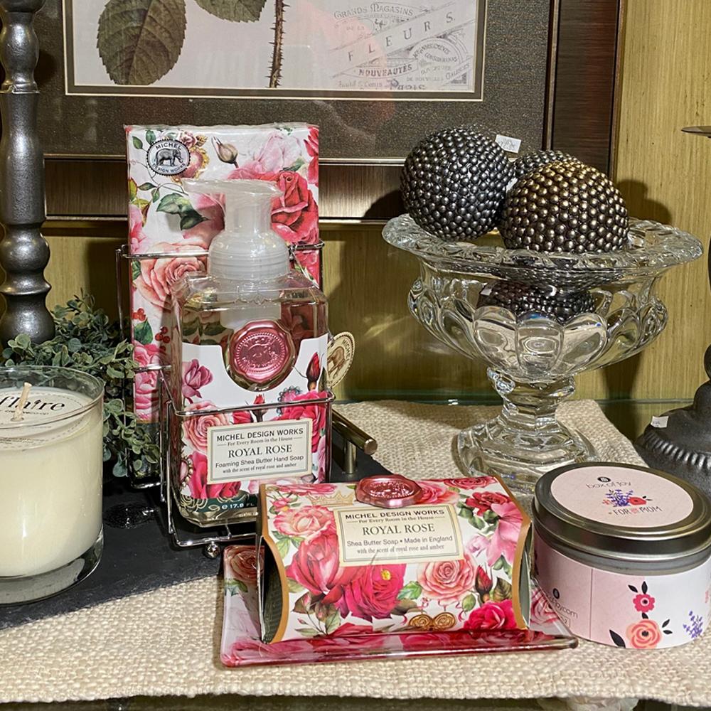 A collection of hand soap, and bar soap wrapped in illustrated red and pink roses, nestled between a candle in a clear vessel and a candle in a closed tin.