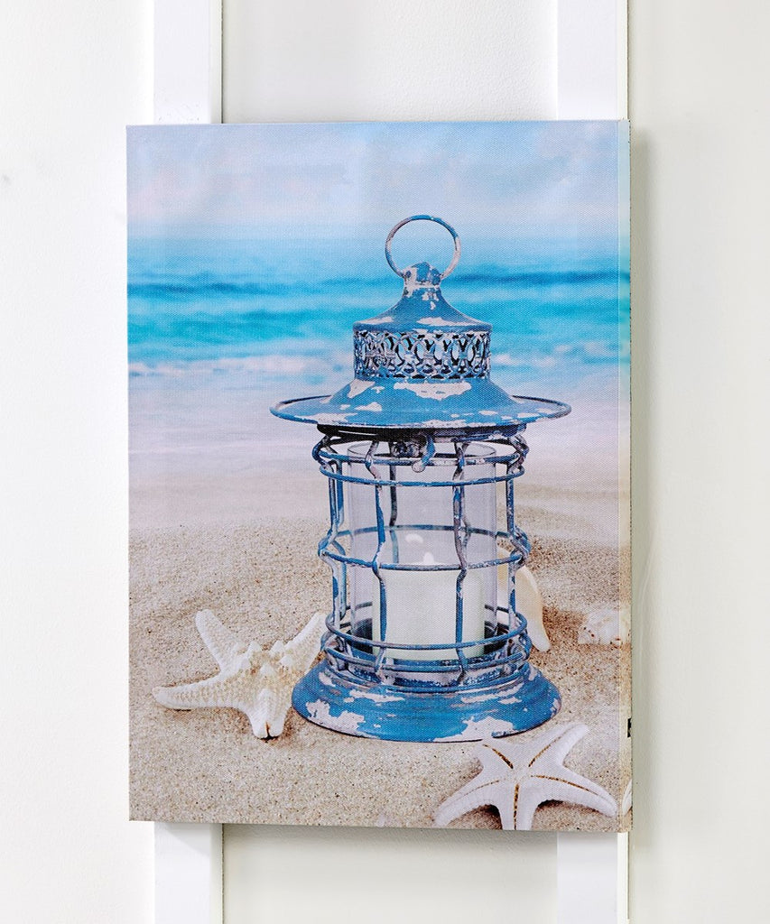 An image of a canvas hung on a white wall.  The canvas has a printed image of a blue weather worn lantern with a white candle in it.  The lantern is sitting in the sand with starfish around it.