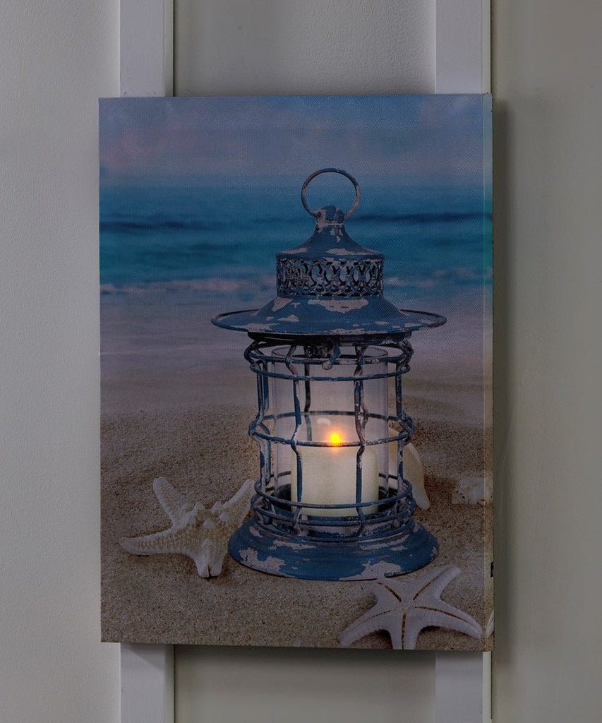 An image of a canvas hung on a white wall.  The canvas has a printed image of a blue weather worn lantern with a white candle in it.  The lantern is sitting in the sand with starfish around it.  This image is darker and shows that there is an orange LED behind the candle on the canvas, it is turned on now and providing a life like glow to the lantern.