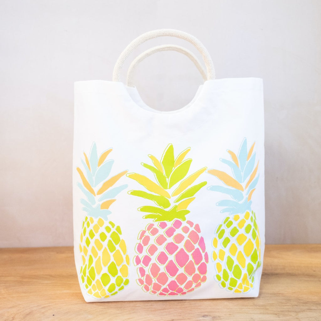 An image of a white tote with arced jute handles on a wooden surface in front of a white wall.  The bag has three pineapples printed on the side in neon colors.  The middle pineapple has leaves of lime green and bright yellow, and the body of it is hot pink and neon orange.  On either side of that pineapple are ones with blue and orange leaves, and yellow and green bodies.