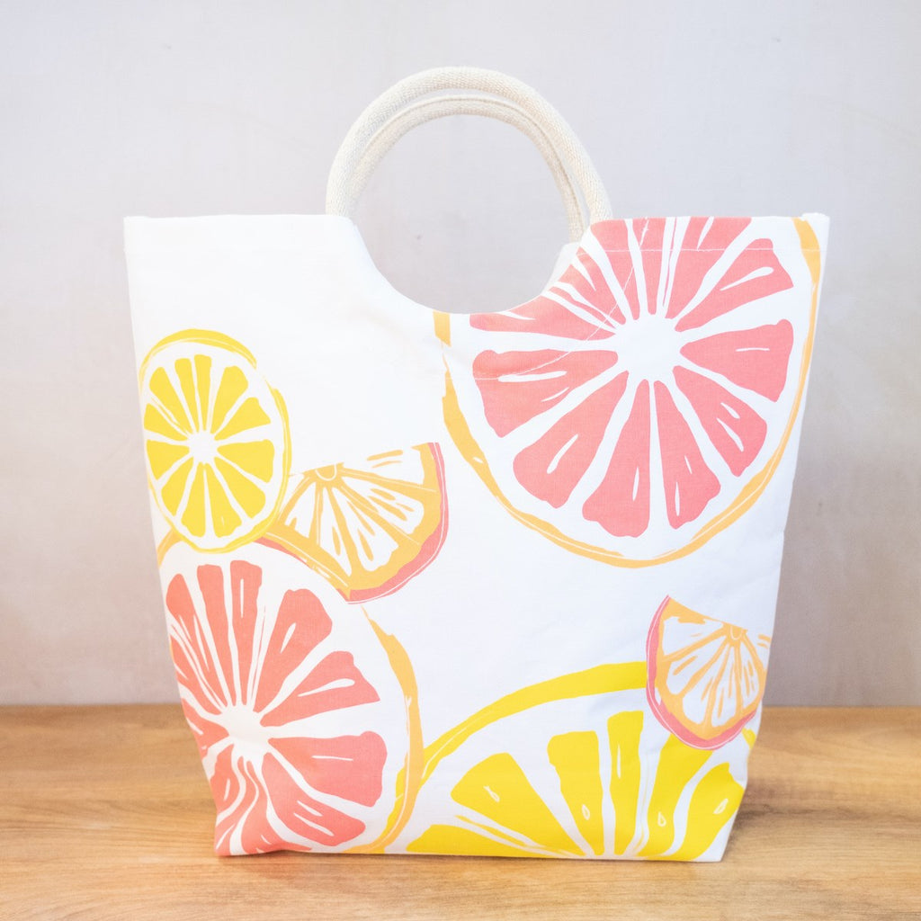 A white tote on a wooden surface in front of a white wall.  The tote has arched handles and a variety of fruit slices printed on the side in yellows, oranges and pinks.