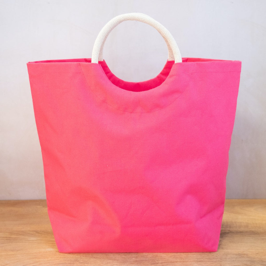 A hot pink tote bag on a wooden surface in front of a white wall.  The bag has arched jute handles and the bag has a semi circle cut below the handle, making a circle of negative space under the handle.