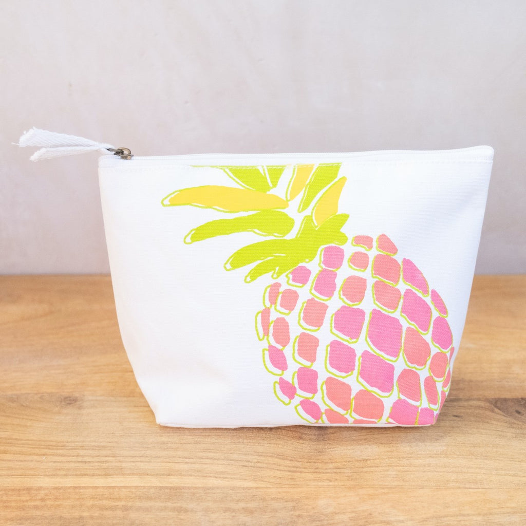 An image of a white cosmetic bag on a wooden surface in front of a white wall.  The cosmetic bag has a zipper on top with two white fabric pulls.  The side of the bag has a neon pineapple printed on it at an angle (so the top and bottom are cut off).  The pineapple has green and yellow leaves and has pink and orange coloration on the body. 
