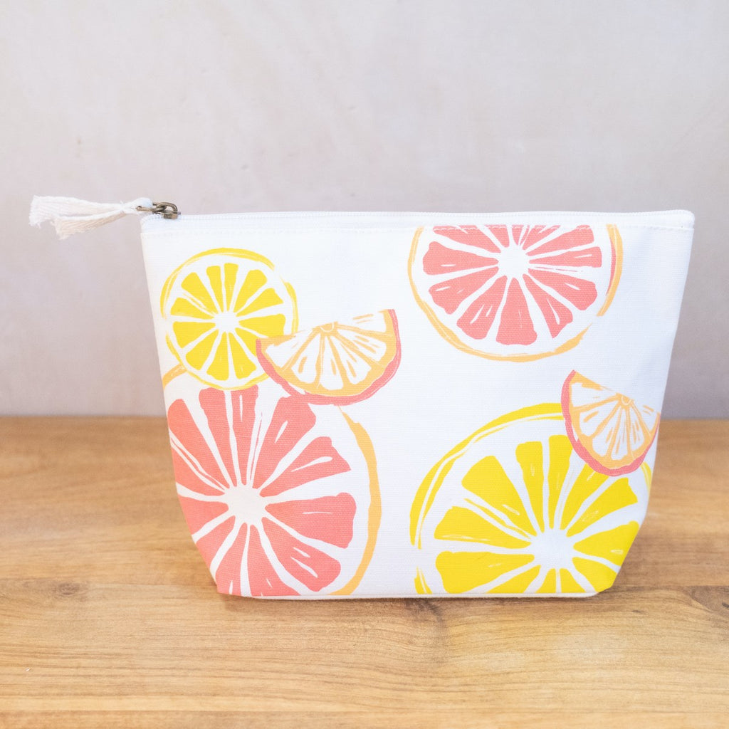 A white cosmetic bag on a wooden surface in front of a white wall.  On the side of the bag are a variety of fruit slices printed on it in orange, yellow, and pink.  The bag has a zipper at the top with two fabric ties.