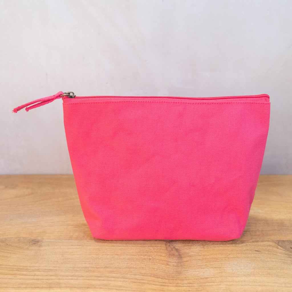 A hot pink cosmetic bag on a wooden surface in front of a white wall.  The bag has a zipper across the top and two pieces of fabric as a pull on the zipper.