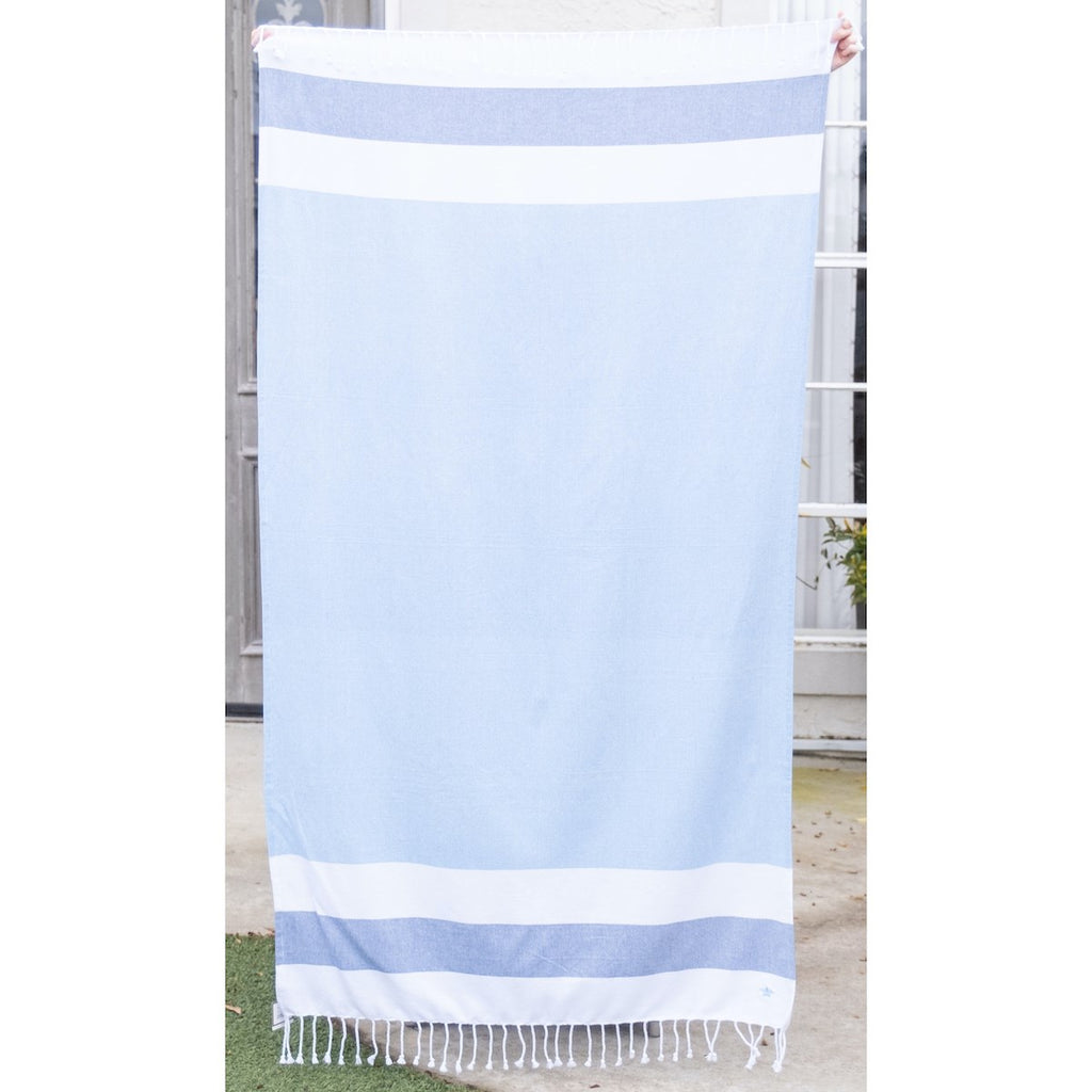 A light blue beach towel with stripes at the top and bottom.  The stripes are wide and horizontal, white, dark blue, white.  There is white fringe along the top and bottom of the towel.