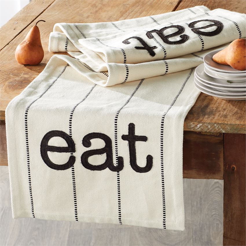 Cotton tan table runner with embroidered lines and appliqué text at each end that says 'eat'.  Runner is folded up at the edge of a wooden table, with one end hanging off.  A stack of plates with a pear on top is on the right, and a single pear is on the left of the runner.