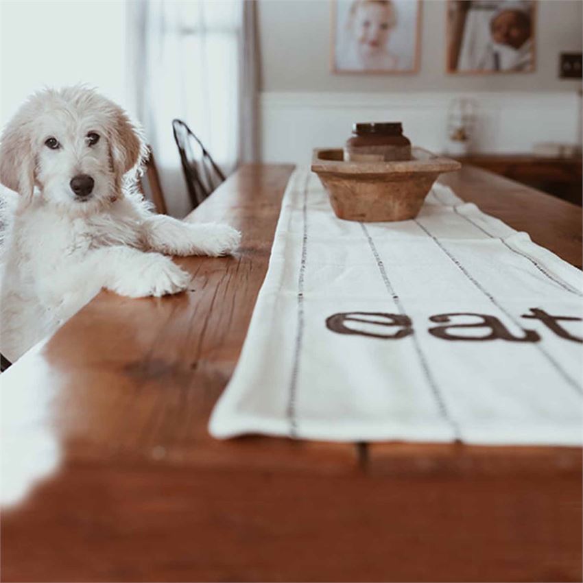 A photo of the table runner on top of a wooden table with a dough bowl on top of the runner.  There is also a white puppy to the left of the runner with its paws on the table.