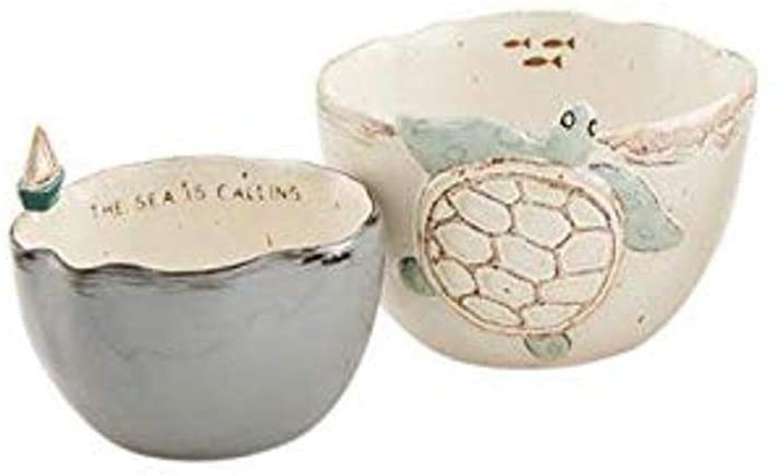 A pair of small bowls on a white background.  The one on the left has a grey outside and dark brown detailing along the wavy rim.  A small ceramic sailboat is permanently attached to the top of the rim.  Just below the rim on the inside, there is text that says 'The Sea is Calling.'  The larger bowl on the right features a dimensional turtle on the outside of the off white bowl.  The turtle has blue/green head and legs.  Just below the rim on the inside are three small brown fish.