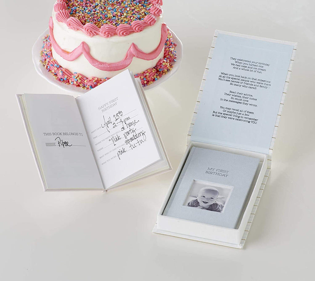 Table with a birthday cake and a guest book for baby opened to an entry page, and a guest book for baby displayed in an open gift box