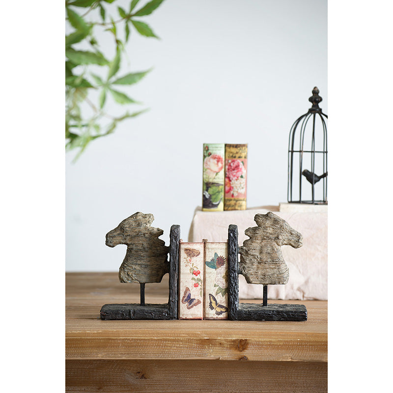 Rustic horse themed bookends.  Horses and supports appear to be carved.  Horses are tan and supports are dark brown.  Bookends are positioned on a table with floral themed books in between.