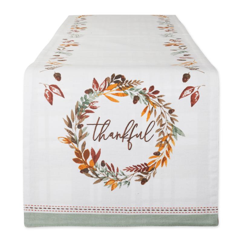 An image of the end of a table runner and the top of it in perspective.  The end of the runner has a bottom border of soft sage green with a stitched border in orange and brown.  Above that is a printed wreath in gold, brown and green, with the word 'thankful' embroidered in brown script in the middle.  The top of the runner features a pattern of various leaves and acorns in the same colors as the wreath.