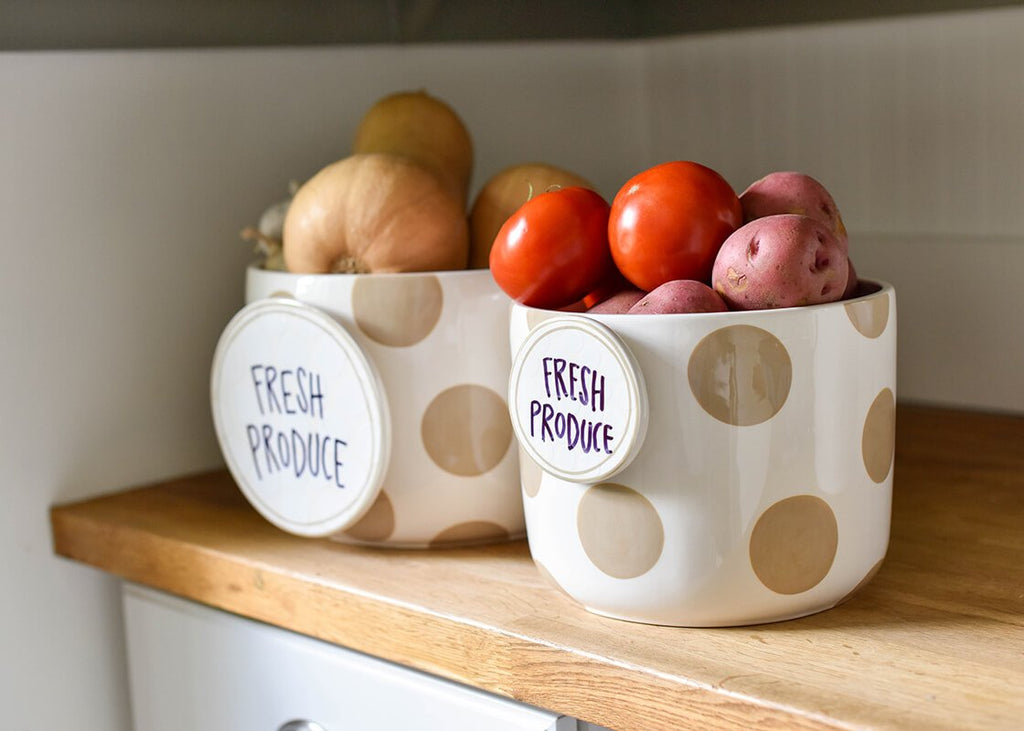 A pair of white bowls with large tan dots on them on a wooden shelf in a white kitchen.  Inside each bowl is a variety of produce (onions, tomatoes, potatoes).  The bowl on the left has a large circular attachment that says 'Fresh Produce' on it.  The bowl on the right has a smaller round disc with the same sentiment, but it is smaller and mounted towards the top of the bowl.