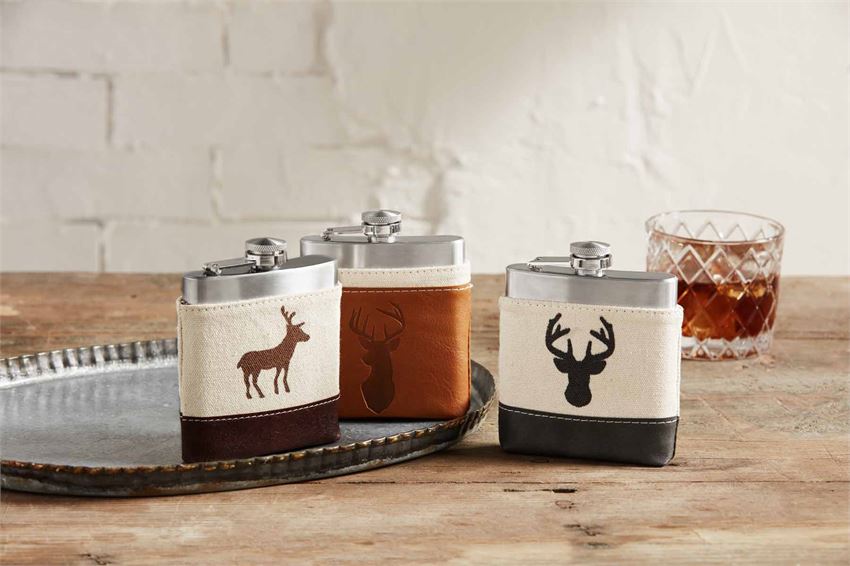 Collection of three flasks resting on a silver serving tray on top of a wooden table.  A rocks glass of whiskey is in the background.  Each stainless steel flask is in a canvas and fake leather pouch with an image of a deer.